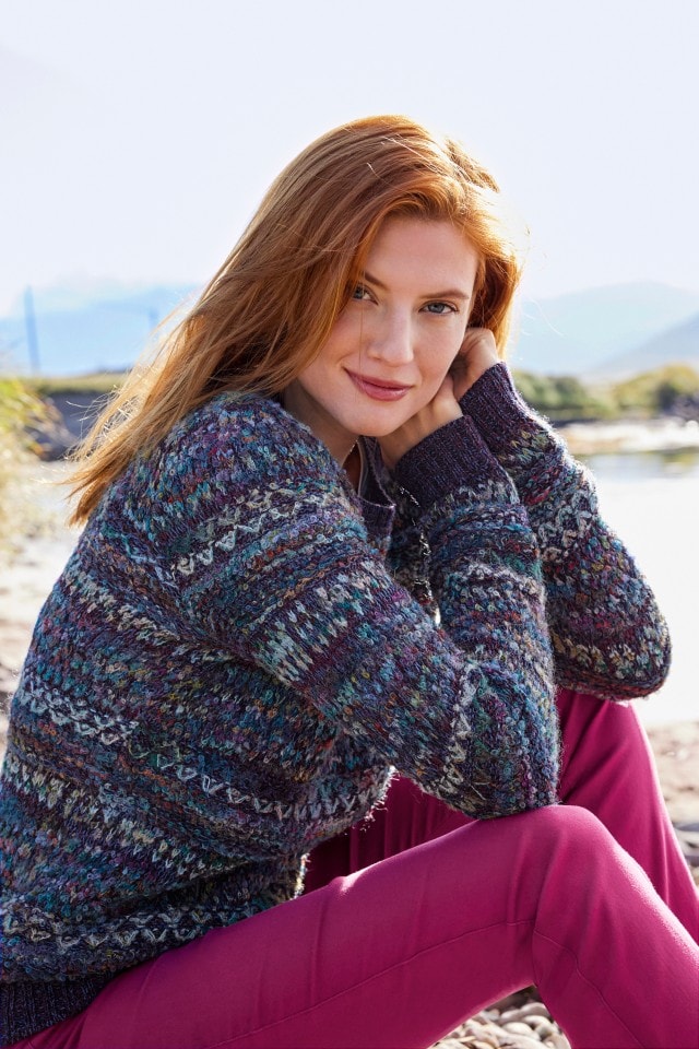 Eine Frau sitzt in einem Alpaka-Pullover in der Sonne am Strand.