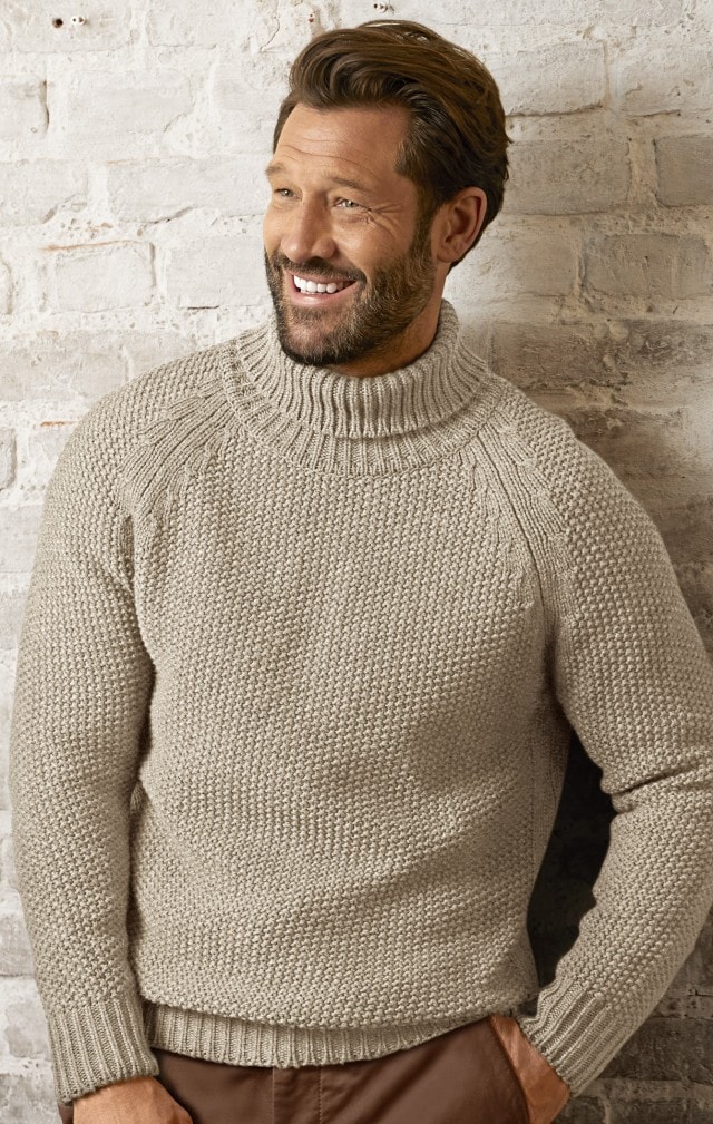 Mann mit beigem Pullover und brauner Hose steht vor einer Wand