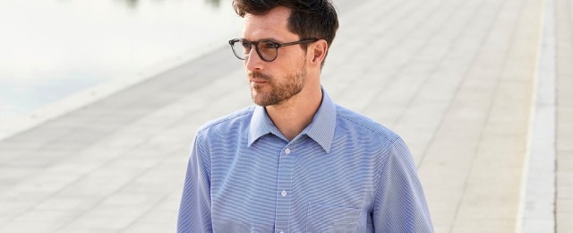 Mann trägt ein hellblau gestreiftes Hemd und eine Brille, während er draußen steht. Der Hintergrund zeigt eine gepflasterte Fläche.