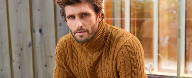 Ein Mann trägt einen grob gestrickten, senfgelben Rollkragenpullover mit Zopfmuster. Er sitzt vor einer Holzwand und einem Fenster.