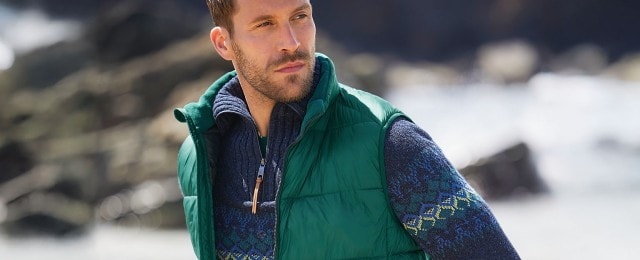 Ein Mann steht draußen und trägt eine grüne Daunenweste über einem blauen Strickpullover mit buntem Muster. Der Hintergrund zeigt eine verschwommene, felsige Landschaft.