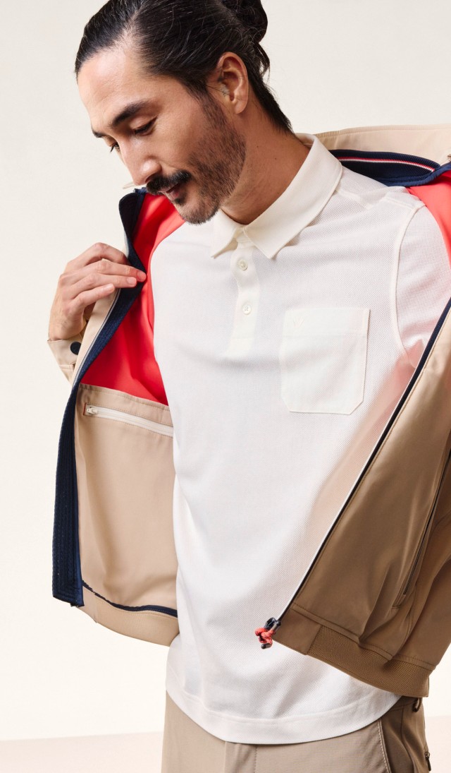 Ein Mann öffnet seine beige Jacke und enthüllt ein weißes Poloshirt darunter. Das Bild vermittelt Stil und Gelassenheit.