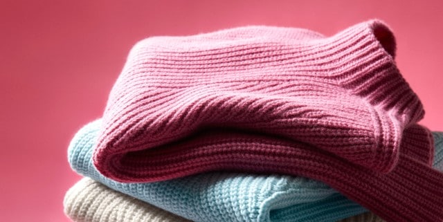 Gestapelte, farbige Cashminopullover in Pink, Blau und Beige vor rosa Hintergrund.