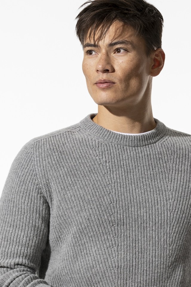 Mann in grauem Strickpullover schaut zur Seite vor hellem Hintergrund.