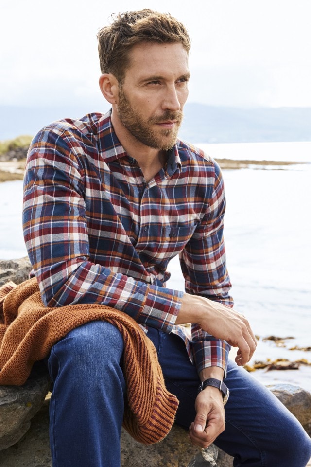 Mann in kariertem Hemd und Jeans sitzt am Ufer, hält einen orangenen Strickpullover.