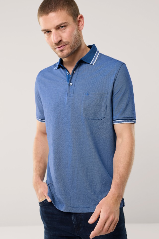 Ein Mann trägt ein blaues, kurzärmliges Poloshirt mit Brusttasche und steht vor einem neutralen Hintergrund.