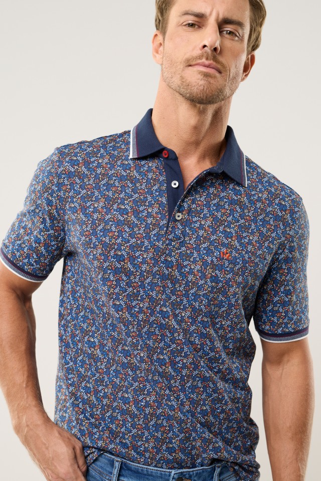 Ein Mann trägt ein kurzärmliges Poloshirt mit kleingeblümtem Muster in Blau- und Rottönen sowie einem dunkelblauen Kragen.