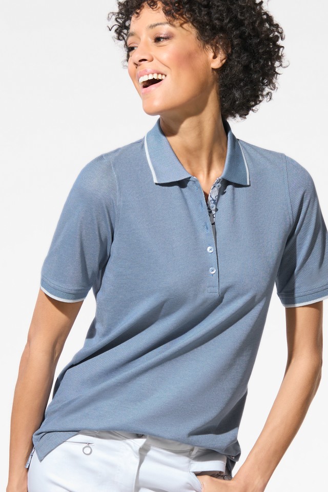 Das Bild zeigt eine Frau, die ein hellblaues Polohemd und eine weiße Hose trägt und zur Seite lächelt.