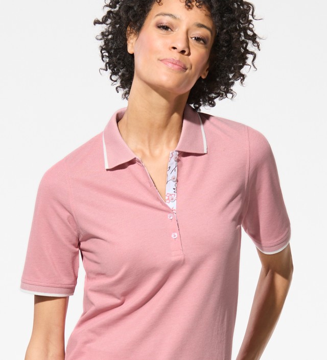 Das Bild zeigt, wie eine Frau in einem rosa Polo-Shirt vor einer Wand steht