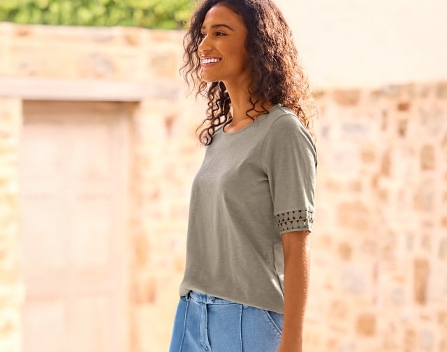 Eine junge Frau mit lockigen Haaren steht lächelnd draußen vor einer hellen Steinmauer und trägt ein Shirt mit Spitzenmuster an den Ärmeln in Khaki sowie eine blaue Hose.
