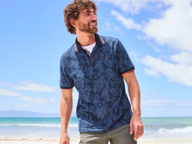 Ein Mann mit lockigen Haaren steht lächelnd am Strand, trägt ein blau gemustertes Poloshirt und im Hintergrund sieht man das Meer und den blauen Himmel mit einigen Wolken.