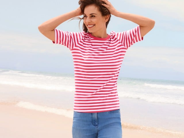 Frau in einem rot-weiß gestreiften T-Shirt und Jeans steht am Strand und schaut zur Seite.
