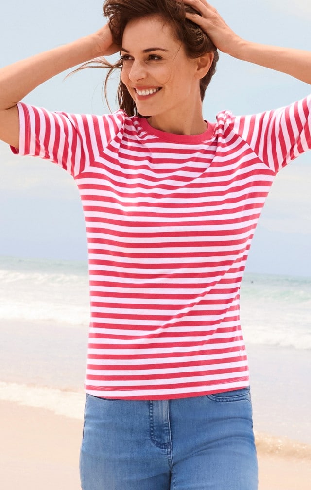 Eine Frau steht am Strand und trägt ein T-Shirt mit roten und weißen Streifen sowie eine blaue Jeans.