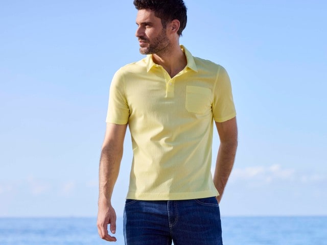 Ein Mann steht am Strand und trägt ein hellgelbes Poloshirt dazu eine Jeans in blau.