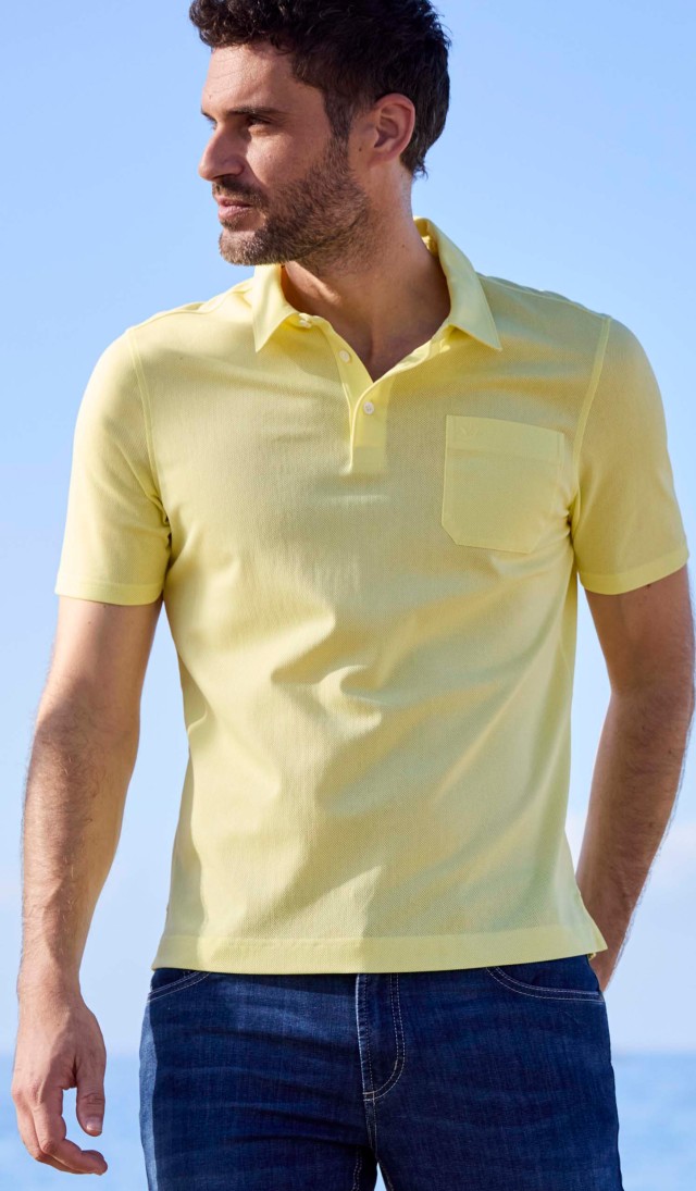 Ein Mann steht am Meer und trägt ein kurzärmliges, hellgelbes Poloshirt mit Kragen und eine dunkelblaue Jeans.