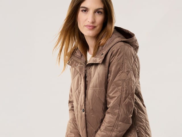 Eine Frau trägt eine gesteppte, braune Kapuzenjacke und blickt ruhig in die Kamera.