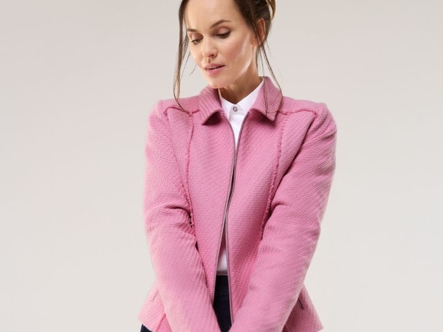 Eine Frau trägt eine rosa Jacke mit Strukturmuster über einer weißen Bluse und blickt nach unten.