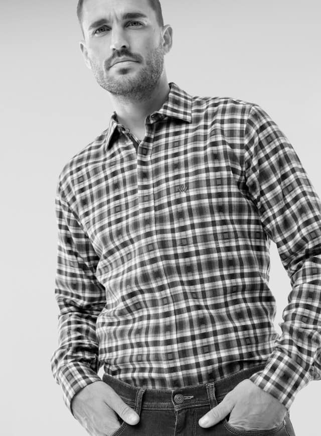 Ein Mann trägt ein kariertes Hemd mit langen Ärmeln und eine dunkle Jeans. Sein Stil wirkt lässig und zugleich gepflegt.