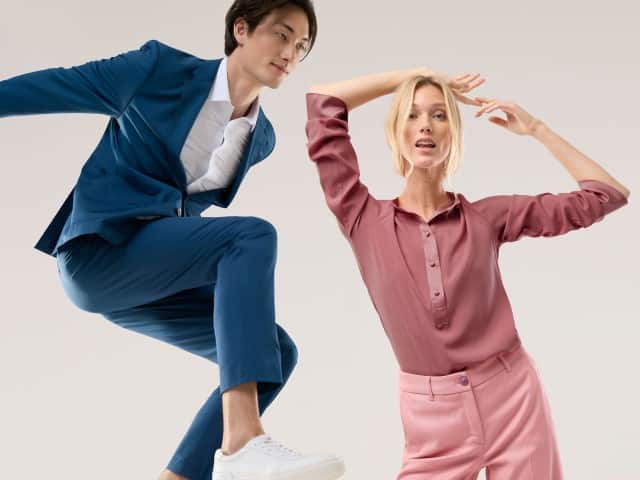 Eine Frau trägt eine Polo Bluse aus Seide in Rosenholz, dazu eine passende Marlenehose. Ein Mann springt in die Höhe und trägt einen Anzug in Marine mit weißem Hemd. 