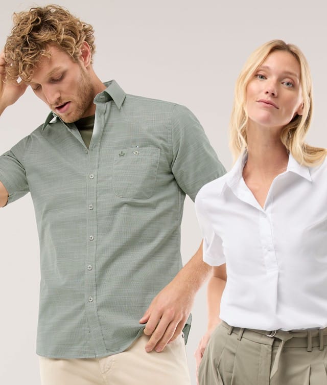 Ein Mann trägt ein Kurzarmhemd in Khaki, eine Frau trägt eine weiße Bluse und hält ihre Hände in den Hosentaschen.