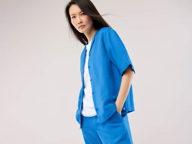 Eine Frau trägt ein leuchtend blaues, kurzärmeliges Set aus Jacke und Hose über einem weißen Oberteil und hat die Hände in den Taschen.