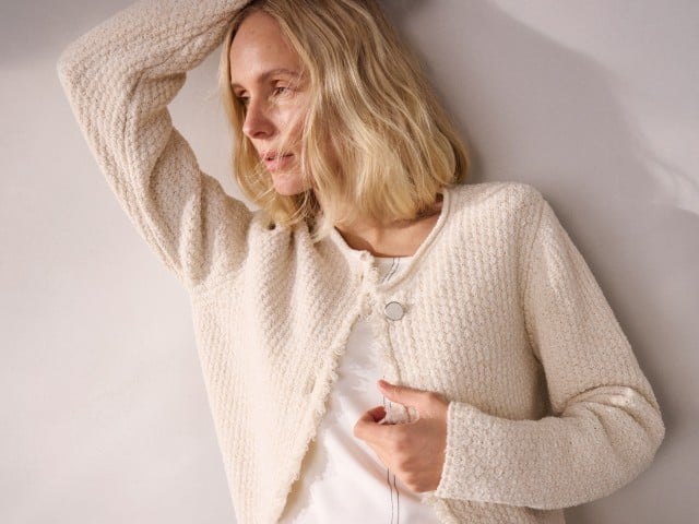 Eine Frau trägt eine cremefarbene, strukturierte Strickjacke über einem hellen Oberteil und lehnt mit erhobenem Arm an einer Wand.