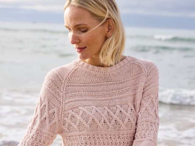 Eine Frau steht am Strand und trägt einen rosafarbenen, grob gestrickten Pullover.