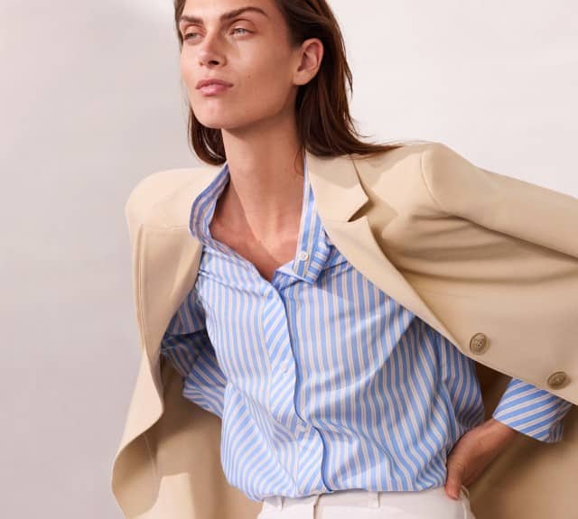 Eine Frau trägt eine beigefarbene Jacke über einer blau‑weiß gestreiften Bluse und blickt leicht zur Seite.