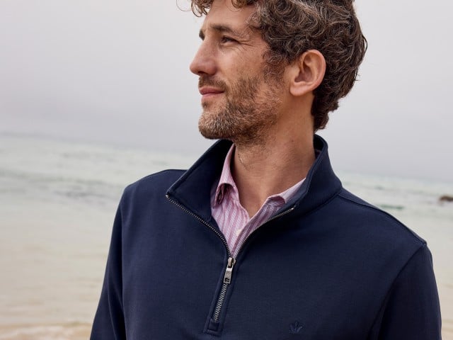 Ein Mann steht am Strand und trägt einen Cardigan in Marine, darunter ein gestreiftes Hemd.