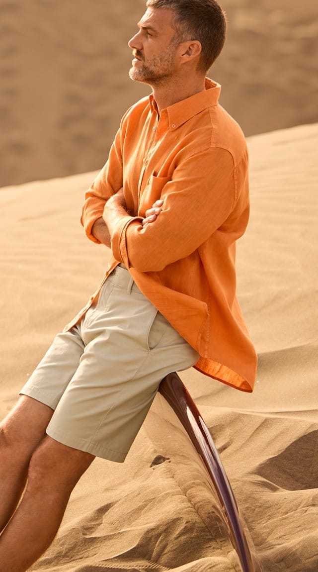 Ein Mann sitzt in einer sandigen Wüstenlandschaft und trägt ein leichtes Sommeroutfit mit einem orangefarbenen Hemd und hellen Shorts, das lässige Eleganz vermittelt.