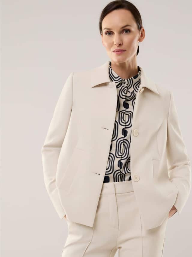 Eine Frau trägt einen eleganten cremefarbenen Hosenanzug mit einer grafisch gemusterten Bluse, das Outfit wirkt ruhig, modern und stilvoll.