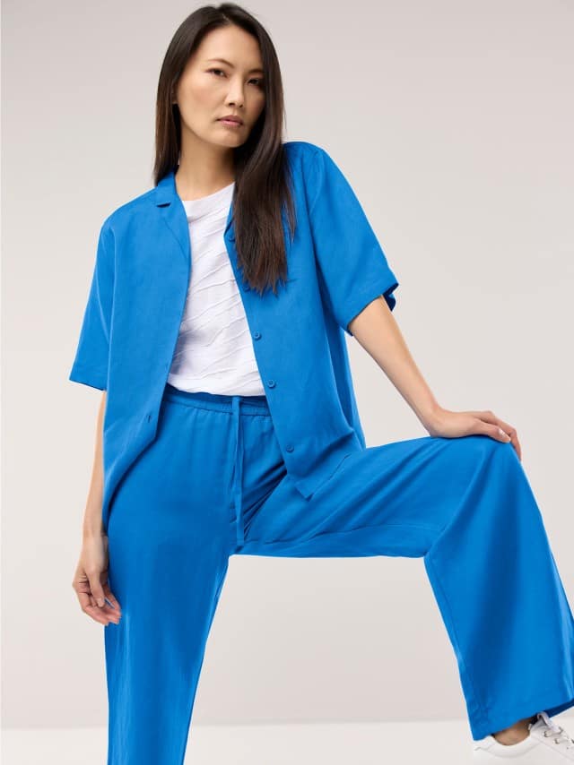 Eine Frau trägt ein leuchtend blaues, locker geschnittenes Set aus Hemd und Hose über einem weißen Oberteil, was einen sportlich‑frischen und energiegeladenen Eindruck vermittelt.