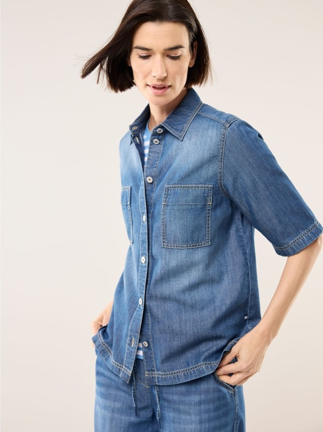 Eine Frau trägt ein lockeres Jeans‑Set aus Hemd und Hose über einem gestreiften Shirt, was einen lässigen, unkomplizierten und entspannt‑frischen Eindruck vermittelt.