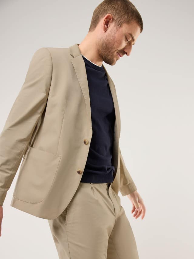 Ein Mann trägt einen beigen, modern geschnittenen Anzug über einem dunkelblauen Shirt, wodurch das Outfit klar, zeitgemäß und entspannt‑stilvoll wirkt.