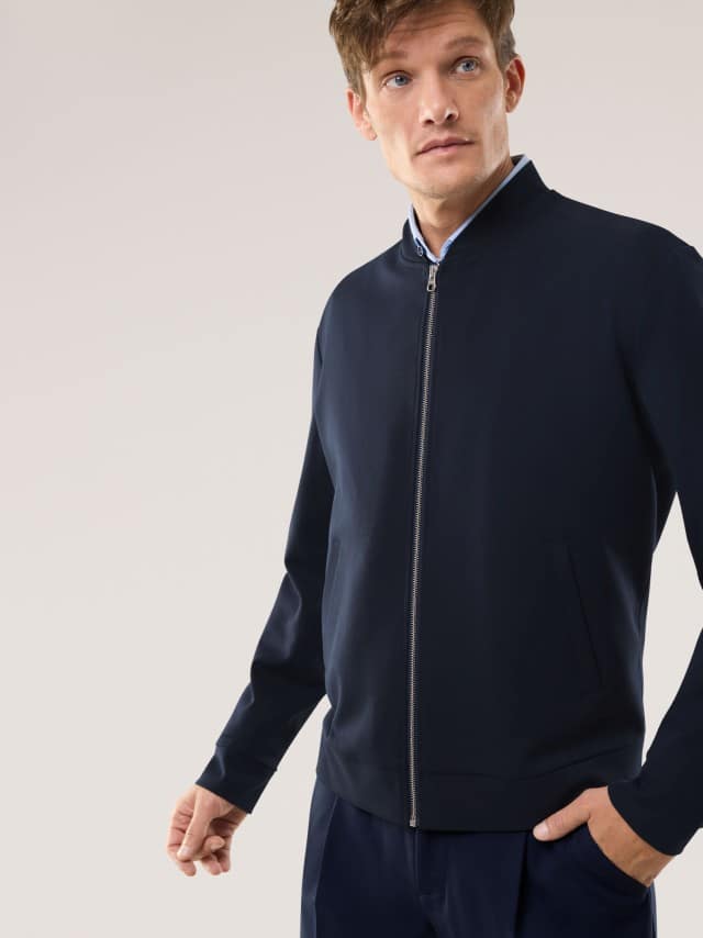 Ein Mann trägt eine dunkelblaue, modern geschnittene Jacke über einem hellen Hemd, was einen klaren, reduzierten und ruhig‑stilvollen Eindruck vermittelt.