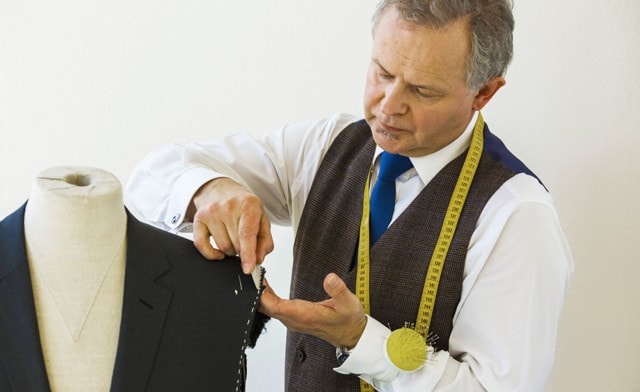 Schneider arbeitet an einer Anzugjacke auf einer Büste, mit Maßband um den Hals.