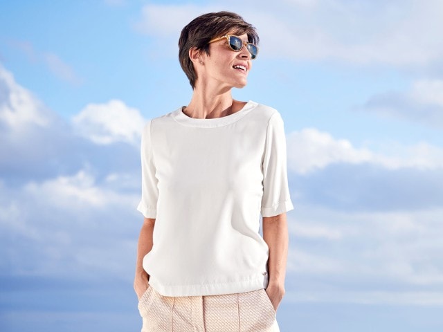Frau mit Sonnebrille und kurzen Haaren trägt eine weiße Shirtbluse, im Hintergrund sind Wolken.