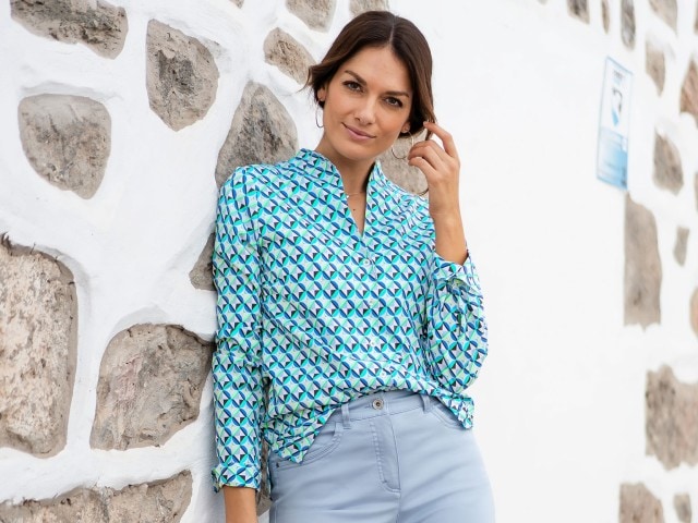 Dame steht vor einer Steinmauer, schaut in die Kamera und trägt eine gemusterte Bluse.