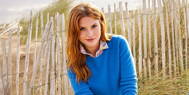 Frau sitzt am Strand, trägt einen blauen Pullover mit einer gemusterten Bluse drunter.