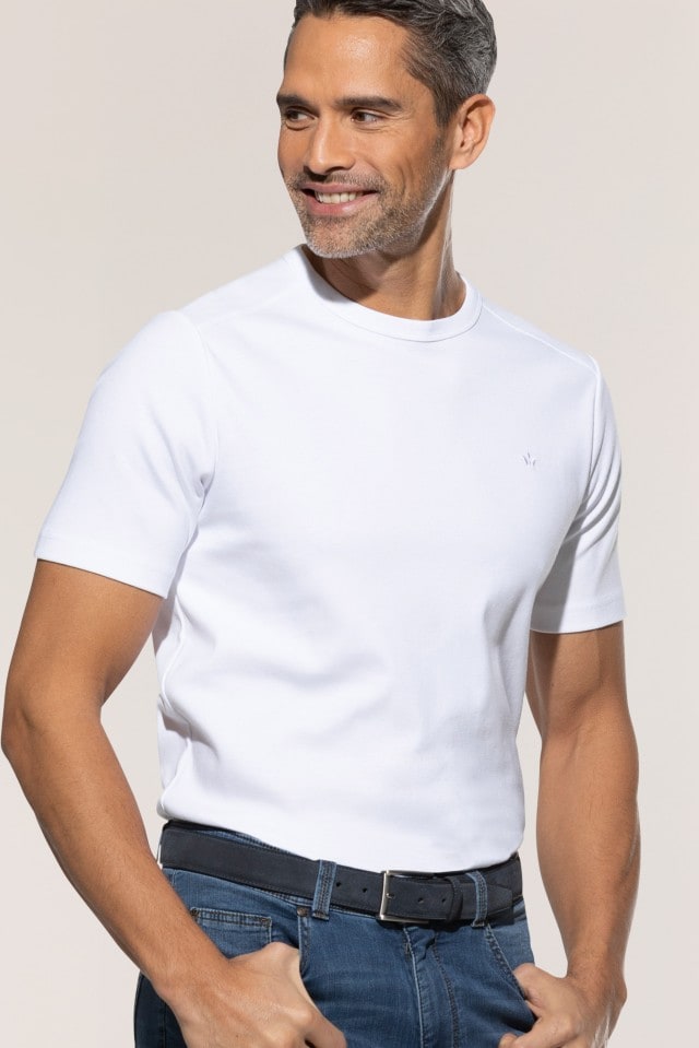 Ein lächelnder Mann trägt ein weißes T-Shirt und eine blaue Jeans mit schwarzem Gürtel vor neutralem Hintergrund.