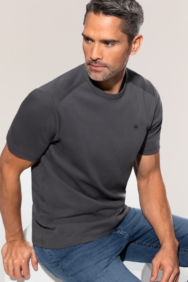 Ein Mann trägt ein dunkelgraues T-Shirt und eine blaue Jeans, während er auf einem weißen Hocker sitzt und zur Seite blickt.