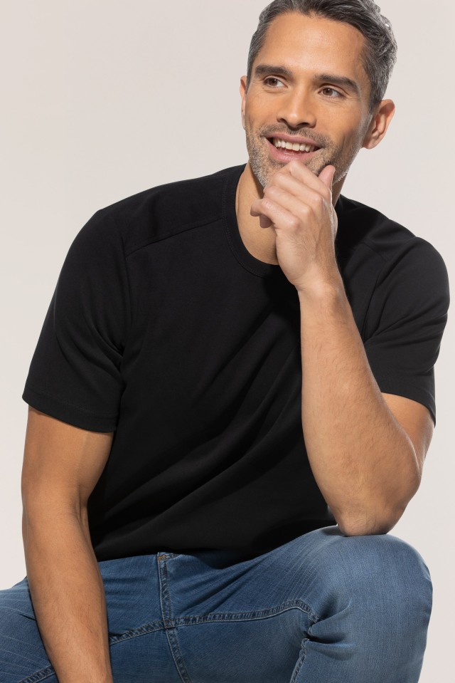 Ein Mann trägt ein schwarzes T-Shirt und blaue Jeans, sitzt entspannt und lächelt, während er die Hand ans Kinn hält.