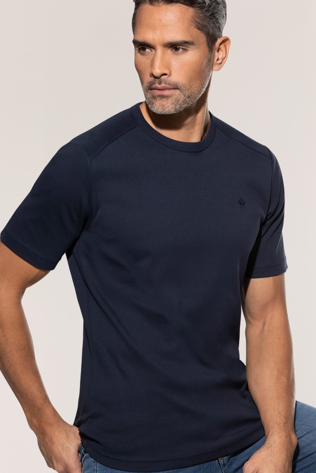 Ein Mann trägt ein dunkelblaues T-Shirt und eine blaue Jeans, während er leicht zur Seite blickt.