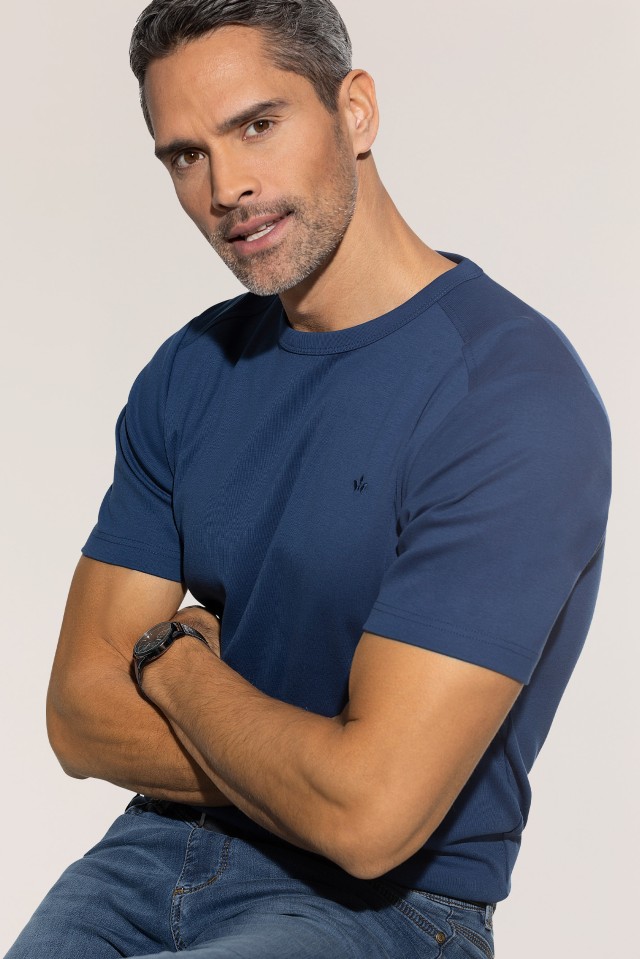 Ein Mann trägt ein blaues T-Shirt und blaue Jeans, hat die Arme verschränkt und lächelt leicht in die Kamera.