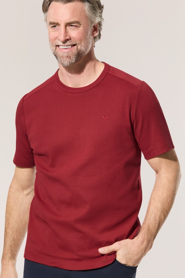 Ein Mann trägt ein bordeauxrotes T-Shirt und dunkle Hosen, lächelt und steckt eine Hand in die Hosentasche.