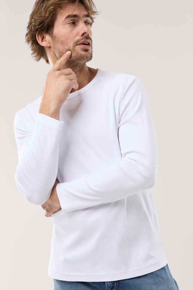 Ein Mann trägt ein weißes Langarmshirt und blaue Jeans, hat eine Hand ins Gesicht gelegt und blickt seitlich nach oben.