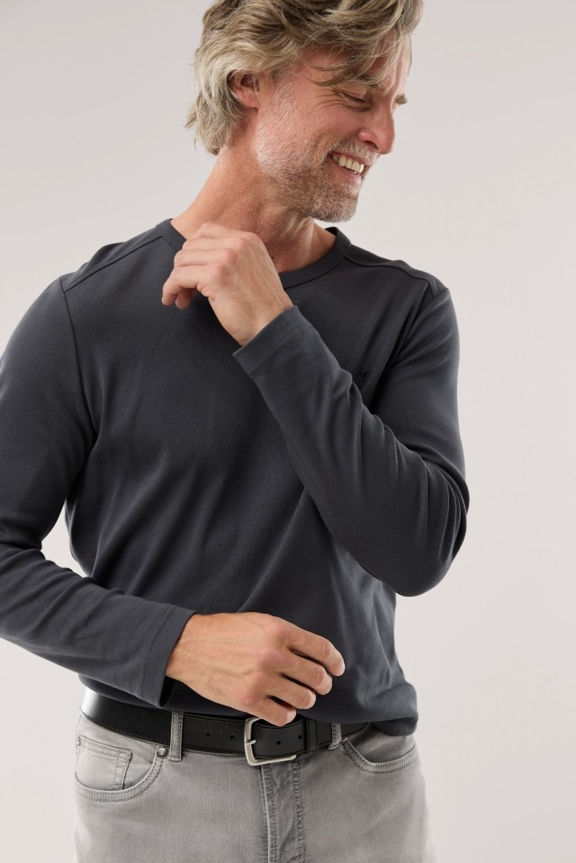 Ein Mann trägt ein dunkelgraues Langarmshirt und eine graue Jeans, lächelt und blickt zur Seite, während er eine Hand zum Kragen hebt.