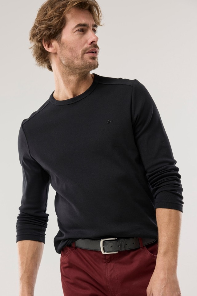 Ein Mann trägt ein schwarzes Langarmshirt und eine bordeauxrote Hose mit schwarzem Gürtel und blickt zur Seite.