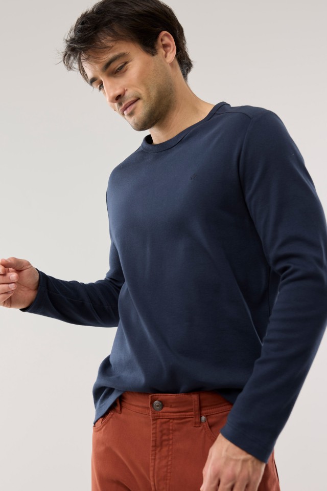 Ein Mann trägt ein dunkelblaues Langarmshirt und eine rostrote Hose, während er mit leicht gesenktem Blick zur Seite schaut.