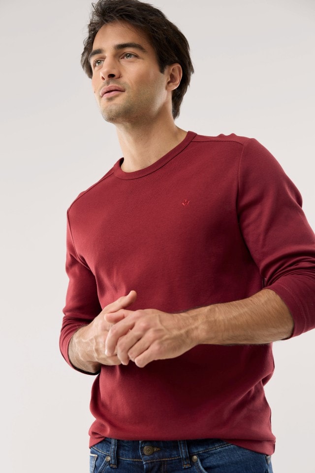 Ein Mann trägt ein bordeauxrotes Langarmshirt und eine blaue Jeans und blickt leicht nach oben.